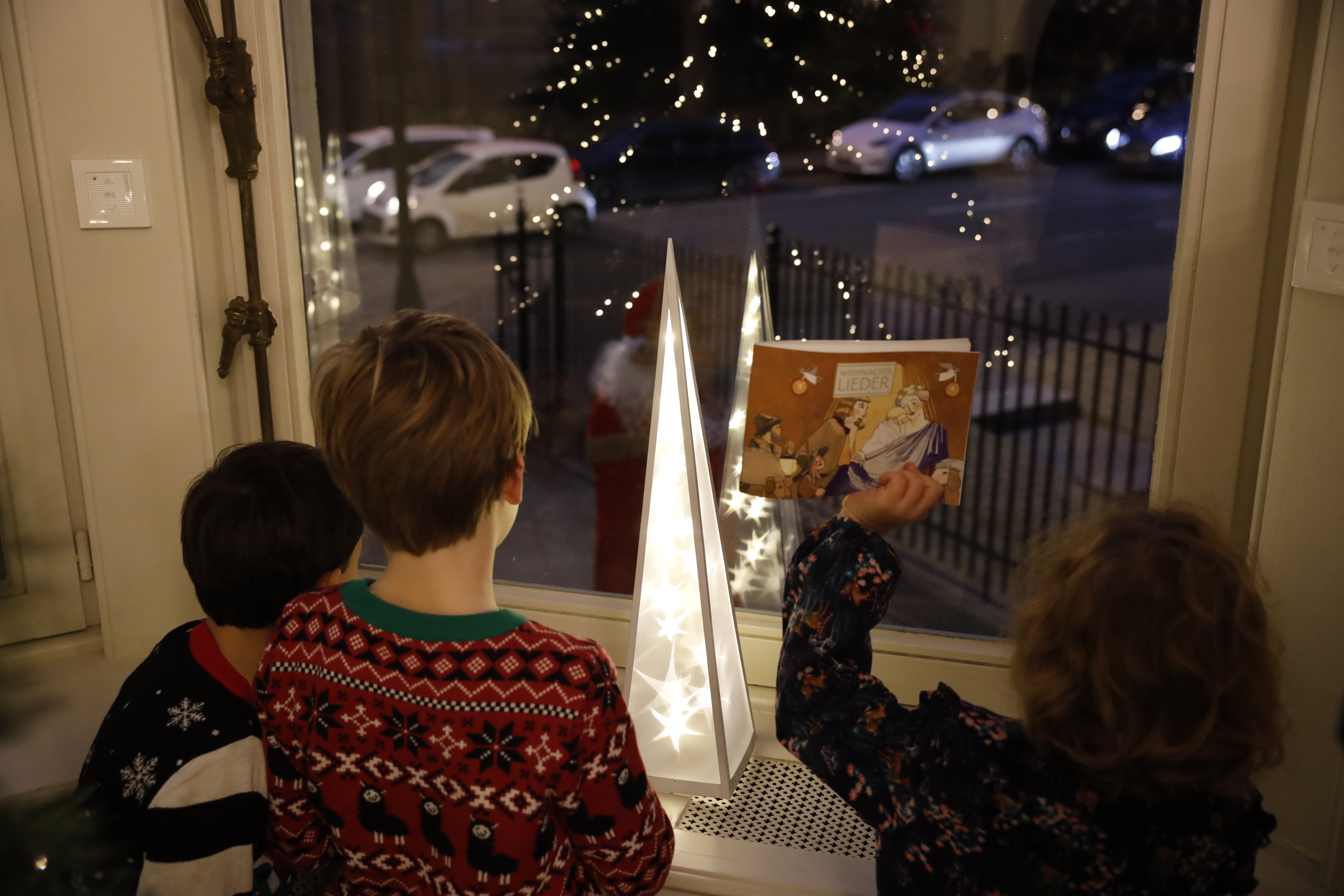 Dieselben drei Kinder stehen vor dem Fenster. Draußen steht de Nikolaus. Ein Mädchen winkt mit einem Liederbuch.