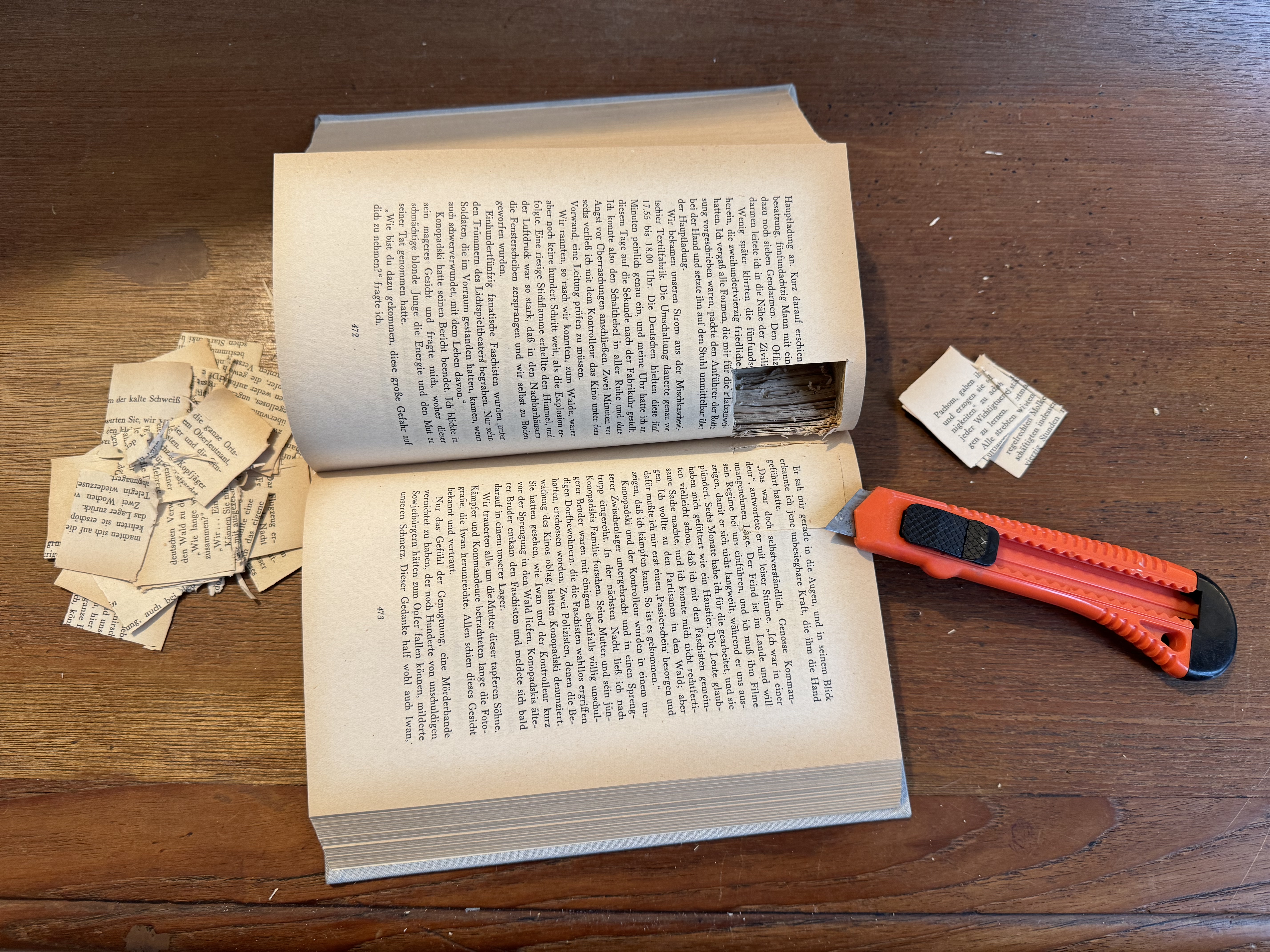 Aufgeklapptes Buch mit einem ausgeschnittenen Versteck für eine Kamera. Daneben ein Teppichmesser und Papierschnippsel