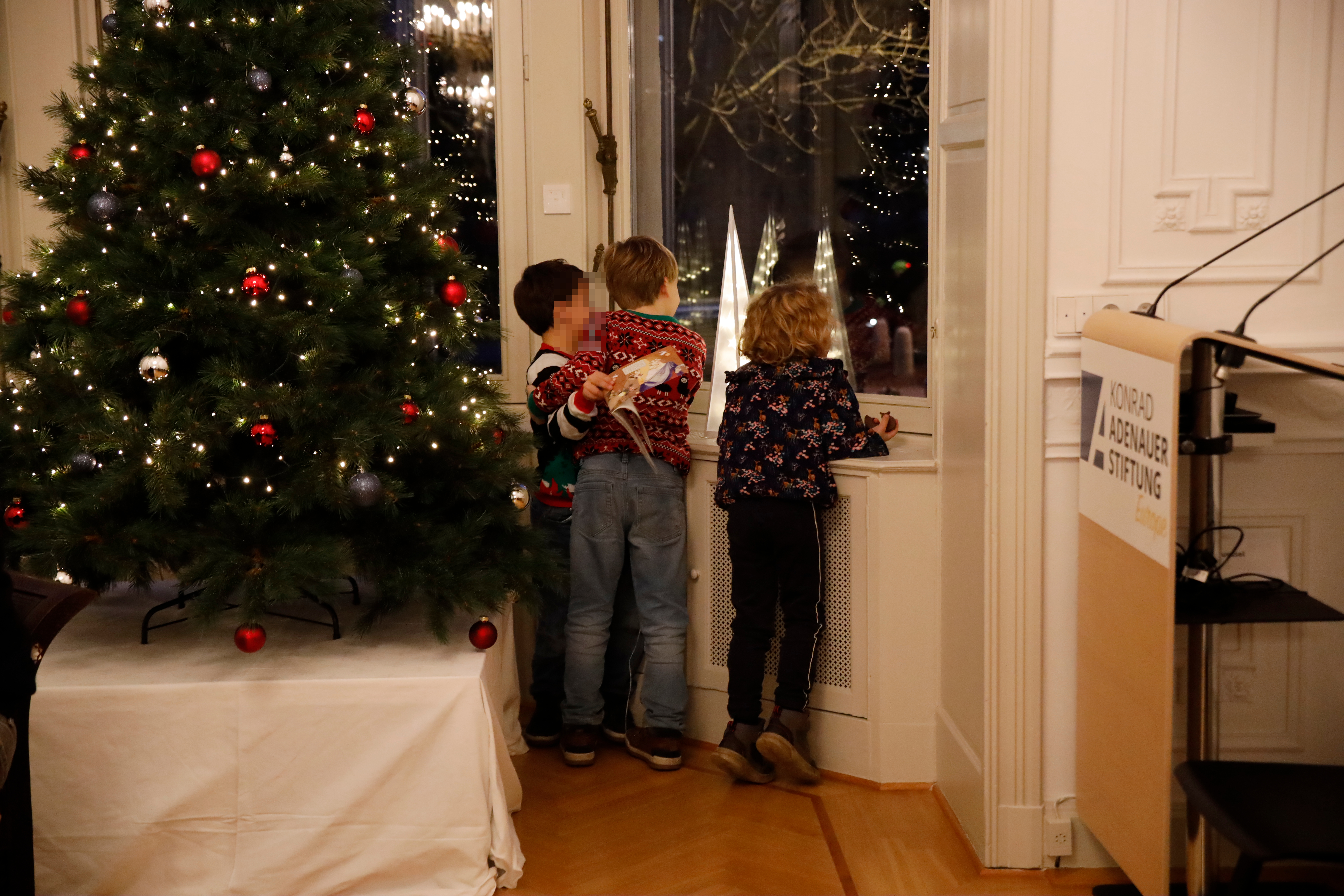 Drei Kinder stehen (mit dem Rücken zur Kamera) erwartungsvoll am Fenster neben dem Weihnachtsbaum. Auf der Fensterbank steht eine spitze Lichpyramide - festliche Stimmung.